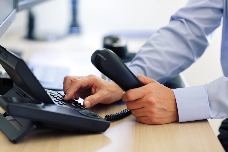 Un professionnel en chemise claire compose un numéro sur un téléphone fixe de bureau dans un environnement de travail moderne