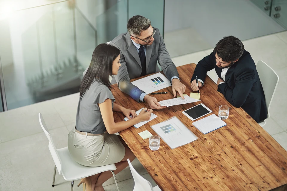 Réunion d'affaires dans bureau moderne avec trois professionnels en costume analysant des rapports financiers autour d'une table en bois
